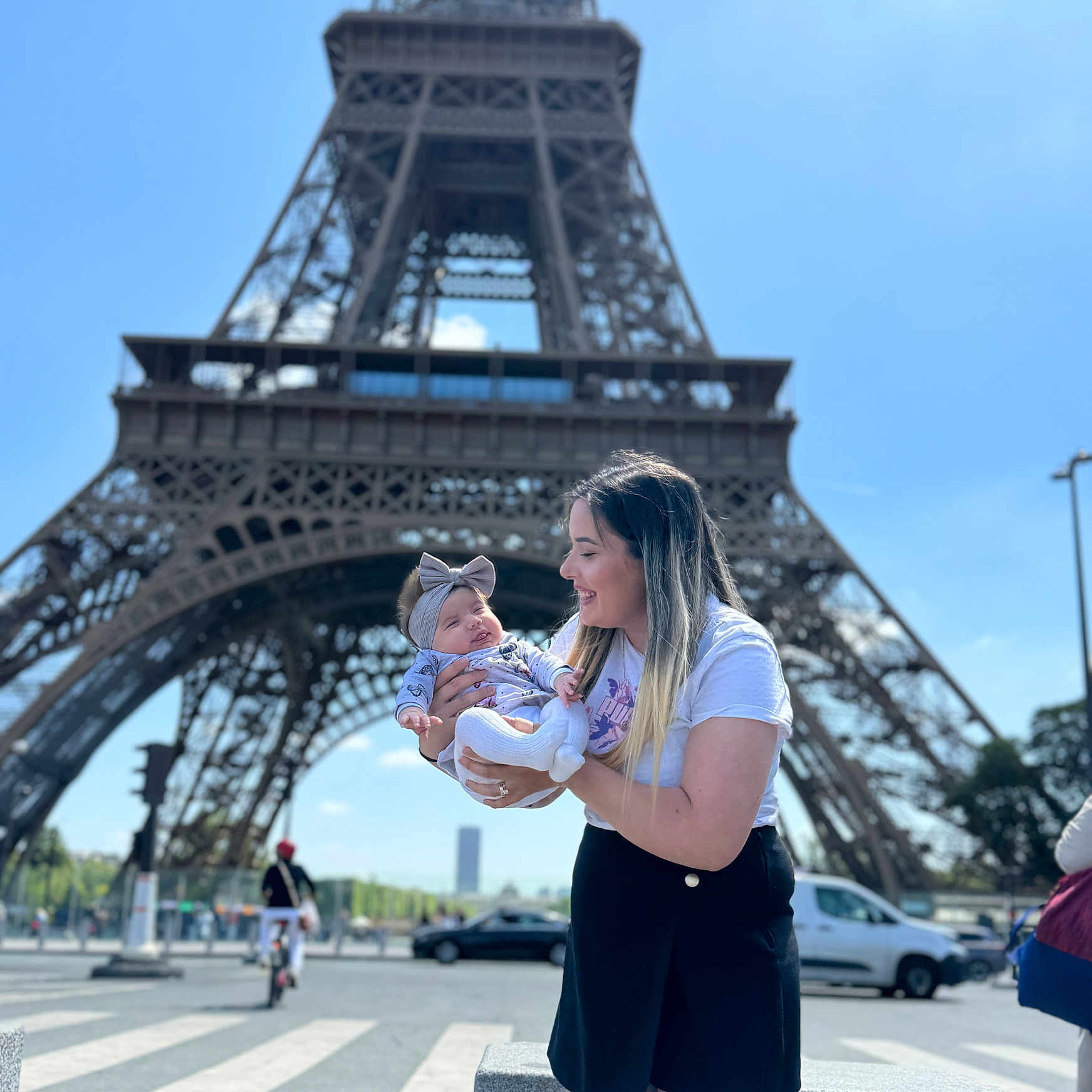 Alma a rejoint le concours — aidez-le/la à gagner de superbes lots ! baby, blue_sky, city, crosswalk, daytime, eiffel_tower, happy, headband, holding, landmark, outdoor, person, skirt, smile, street, tourist, traffic, tshirt, urban, woman