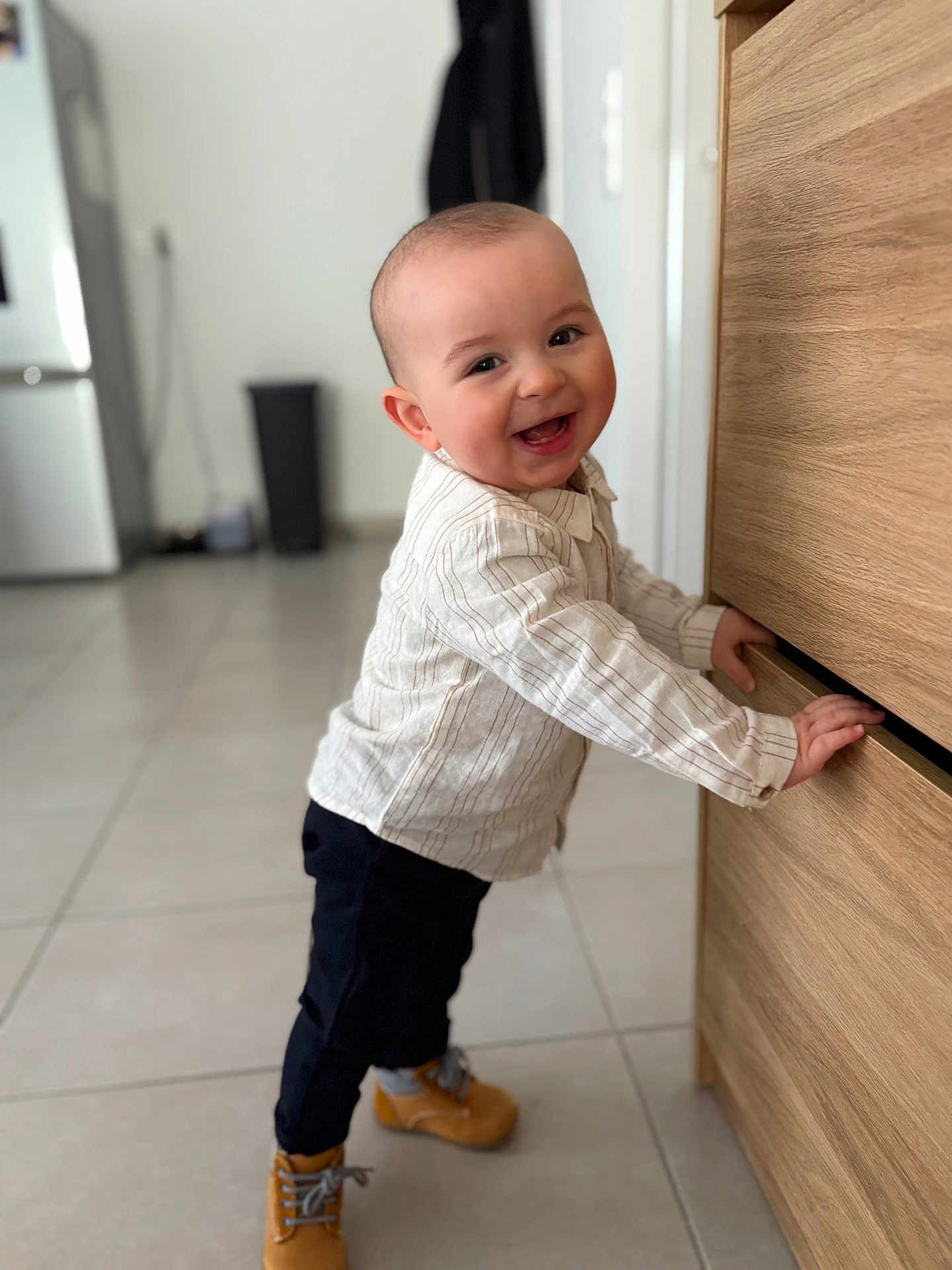 Vittorio participe au concours pour gagner de l'argent avec cette photo : baby, toddler, smiling, standing, wooden_drawer, kitchen, boots, striped_shirt, pants, tile_floor, happy, face, hands, eyes, furniture, home_interior, refrigerator, trash_bin, floor_tiles, portrait