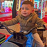 amusement, arcade, boy, child, colorful, entertainment, floor, fun, game_machine, hands_on_handlebars, indoor, jacket, lighting, motorcycle_ride, neon_lights, pants, playful, smiling, sneakers, young