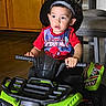 toddler, child, firefighter_hat, spiderman_shirt, toy_atv, indoor, wooden_cabinet, chair, floor, curious, hands, face, eyes, expression, clothing, footwear, play, ride, vehicle, young_child
