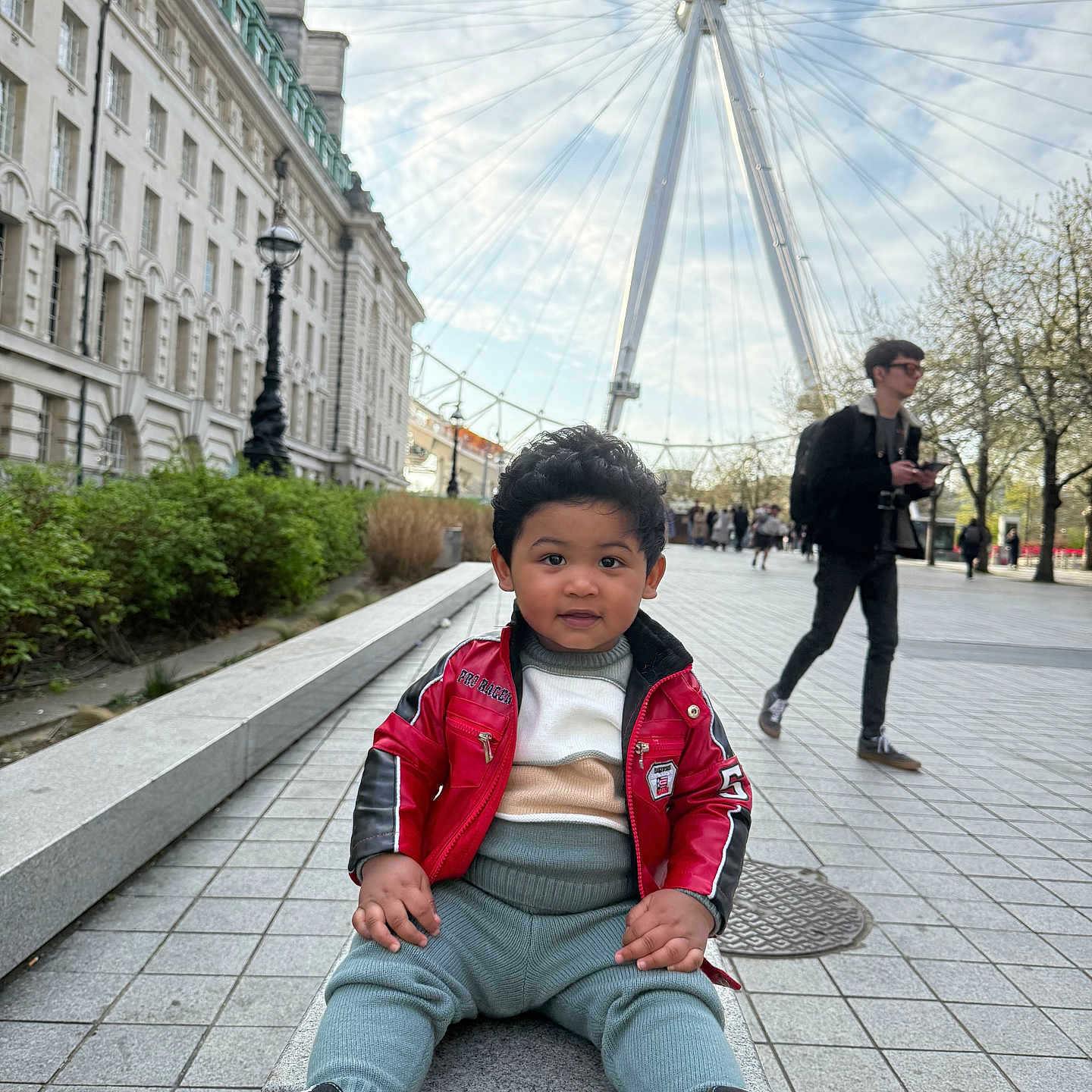 Zaviyaan is registered to the contest to win money with this photo: bench, building, casual_clothing, child, cityscape, curly_hair, daytime, ferris_wheel, outdoor, people, plaza, red_jacket, sidewalk, sky, smiling, sneakers, toddler, trees, urban, walking
