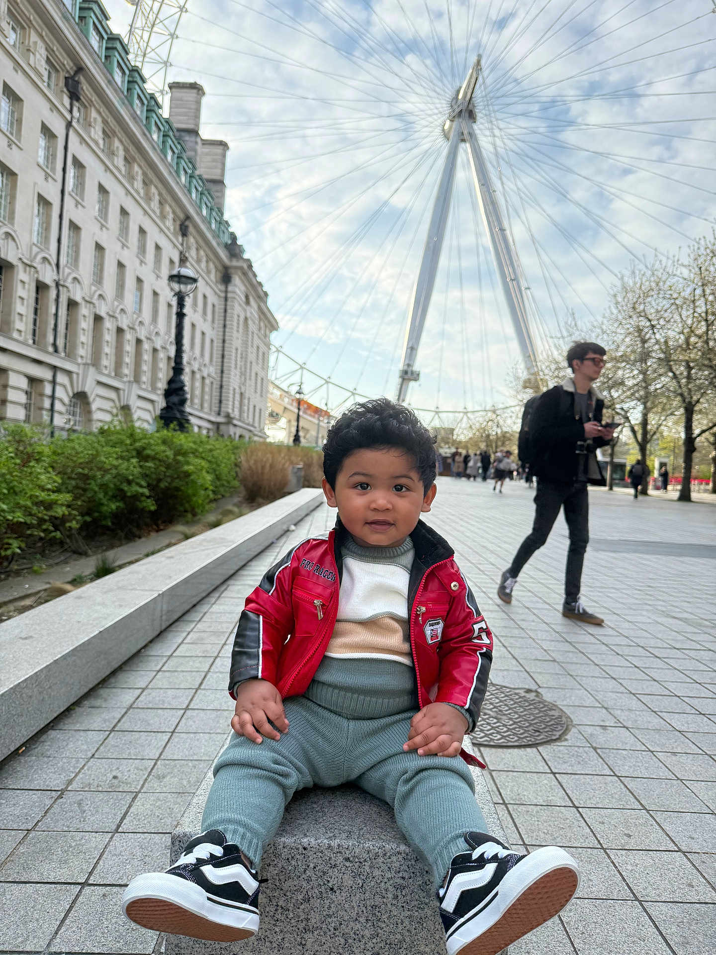 Zaviyaan is registered to the contest to win money with this photo: bench, building, casual_clothing, child, cityscape, curly_hair, daytime, ferris_wheel, outdoor, people, plaza, red_jacket, sidewalk, sky, smiling, sneakers, toddler, trees, urban, walking