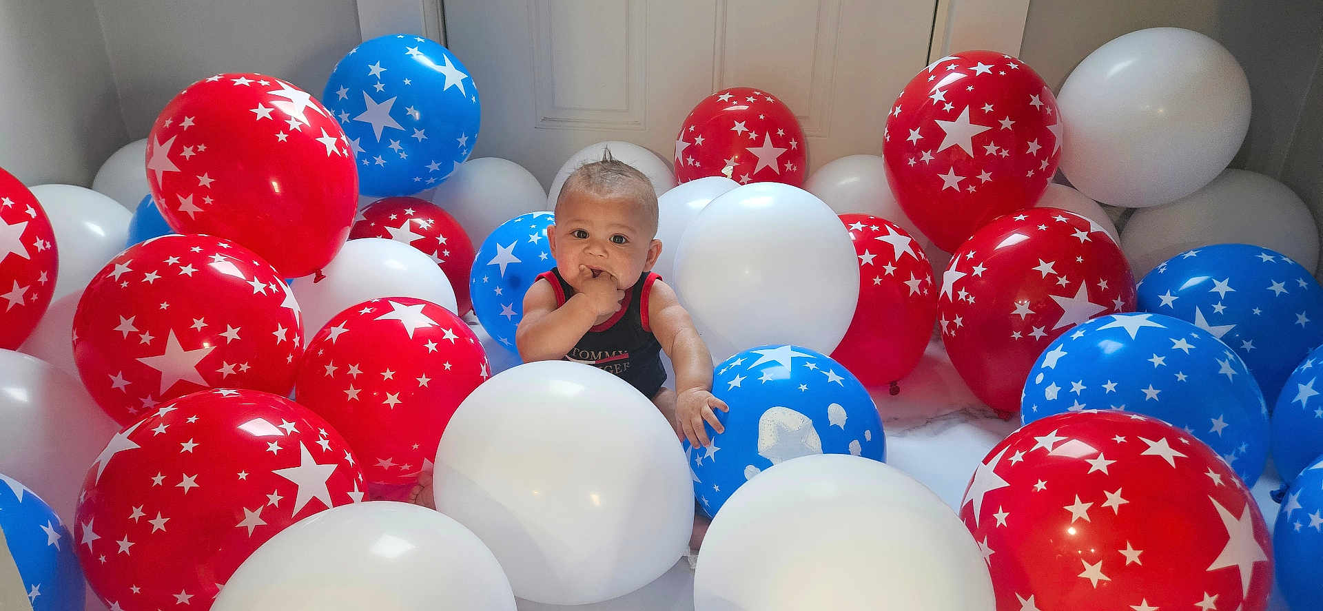 Zavion is registered to the contest to win money with this photo: baby, balloons, red_balloons, blue_balloons, white_balloons, stars, indoors, curious, child, portrait, celebration, party, colorful, cute, infant, sitting, hand_in_mouth, mohawk_hair, floor, decoration