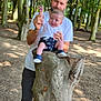 baby, man, tree_stump, forest, outdoor, nature, trees, smiling, holding, casual_clothing, shorts, sneakers, greenery, sunlight, daylight, happy, person, child, parent, family