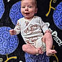 baby, infant, onesie, smile, face, blanket, pattern, purple, black, cartoon, text, lying_down, cute, child, happy, toddler, clothing, playful, young, indoors