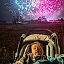 baby, stroller, fireworks, night, hoodie, toy, blanket, outdoor, child, field, colorful, celebration, lights, darkness, grass, person, seat, festive, sky, event
