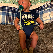 Jamie is registered to the contest to win money with this photo: baby, infant, pacifier, onesie, clothing, blanket, pillow, bed, socks, lying_down, child, cute, indoors, soft_texture, patterned, blue, yellow_text, expression, face, person