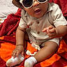 baby, child, sunglasses, hat, beige_outfit, socks, blanket, red_blanket, orange_blanket, indoor, curly_hair, pacifier, baby_clothing, cute, playful, seated, infant, portrait, person, cozy