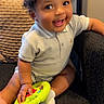 baby, child, curly_hair, smiling, toy, indoor, chair, sitting, happy, cute, person, sock, short_sleeve_shirt, playful, bright_eyes, young_child, toddler, hand, furniture, portrait