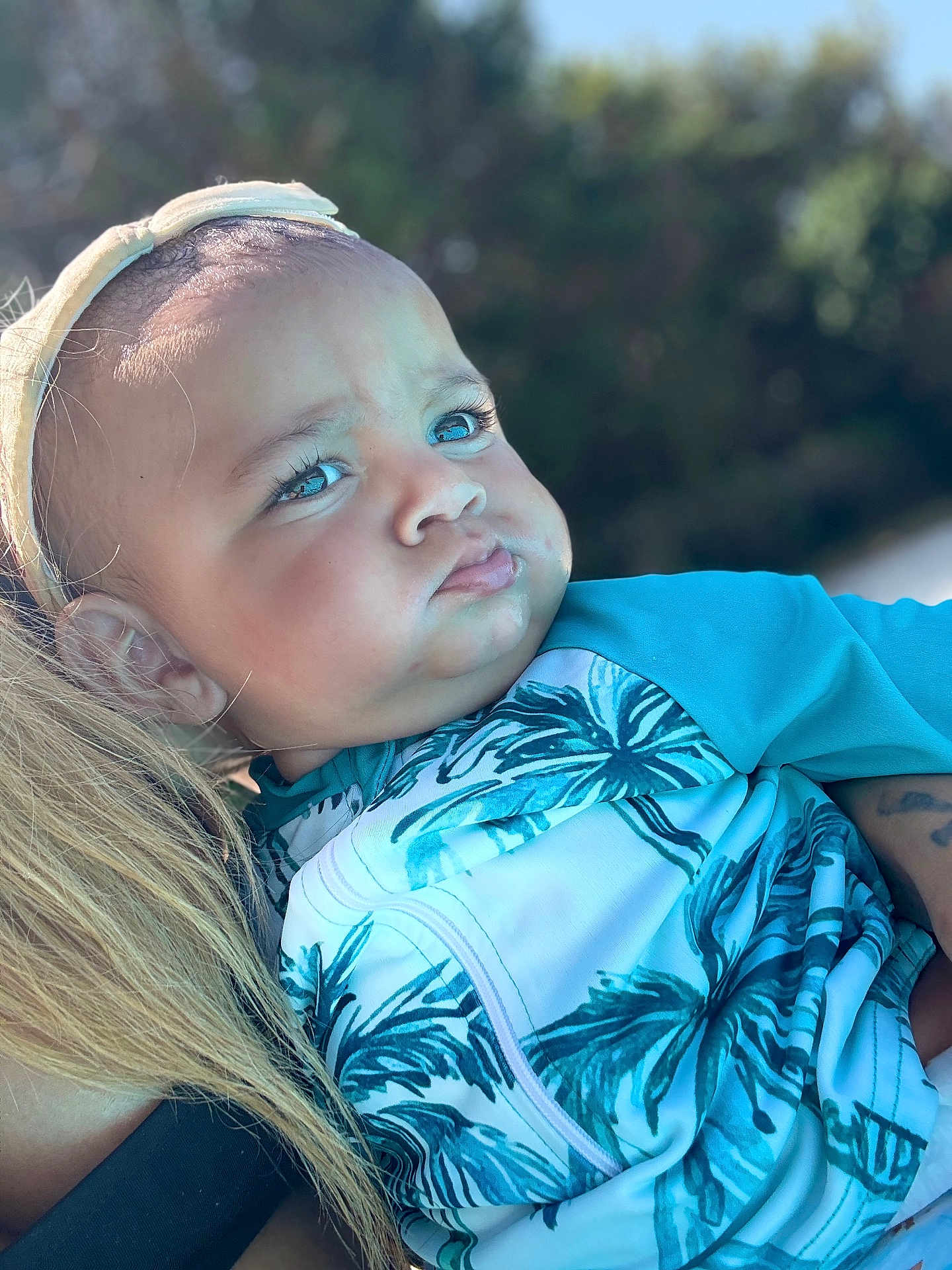 Imran a rejoint le concours — aidez-le/la à gagner de superbes lots ! baby, child, face, blue_eyes, headband, leaf_pattern, clothing, person, outdoor, nature, portrait, expression, holding, blonde_hair, skin, cute, serious, young_child, close_up, sunlight