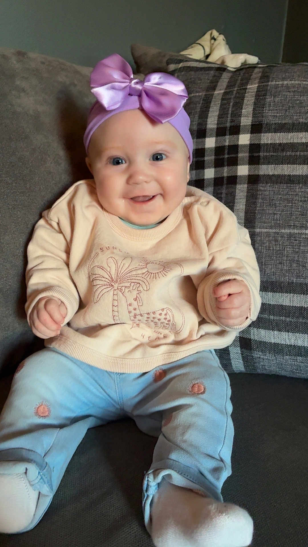 Lottie-Mae is registered to the contest to win money with this photo: baby, child, smiling, headband, bow, sweater, pants, socks, couch, cushion, plaid, indoor, happy, seated, portrait, infant, cute, cozy, clothing, person