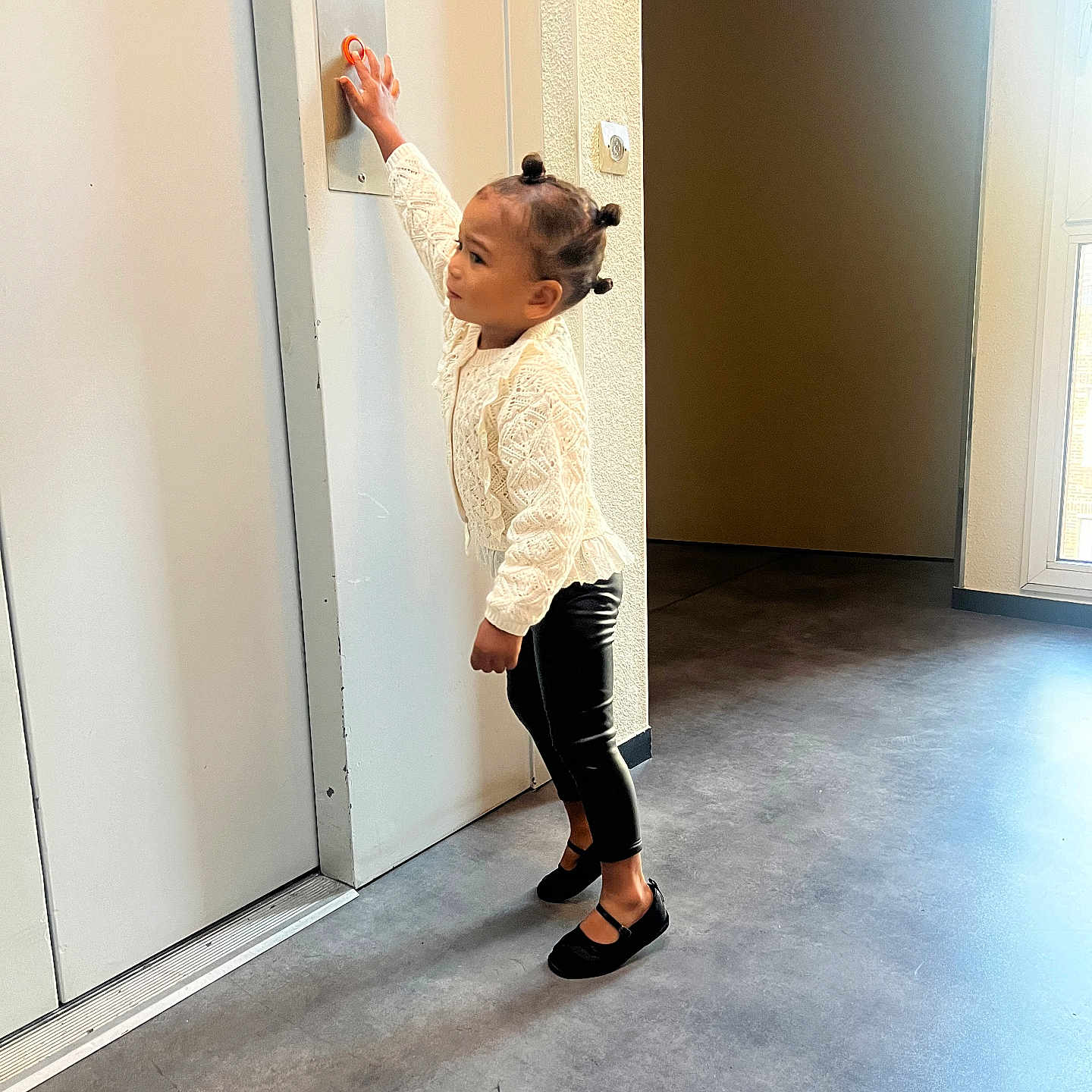 Layna participe au concours pour gagner de l'argent avec cette photo : child, indoor, door, button, floor, light, window, hair_buns, white_sweater, black_pants, shoes, curious, standing, young_child, wall, panel, natural_light, concrete_floor, person, casual_clothing