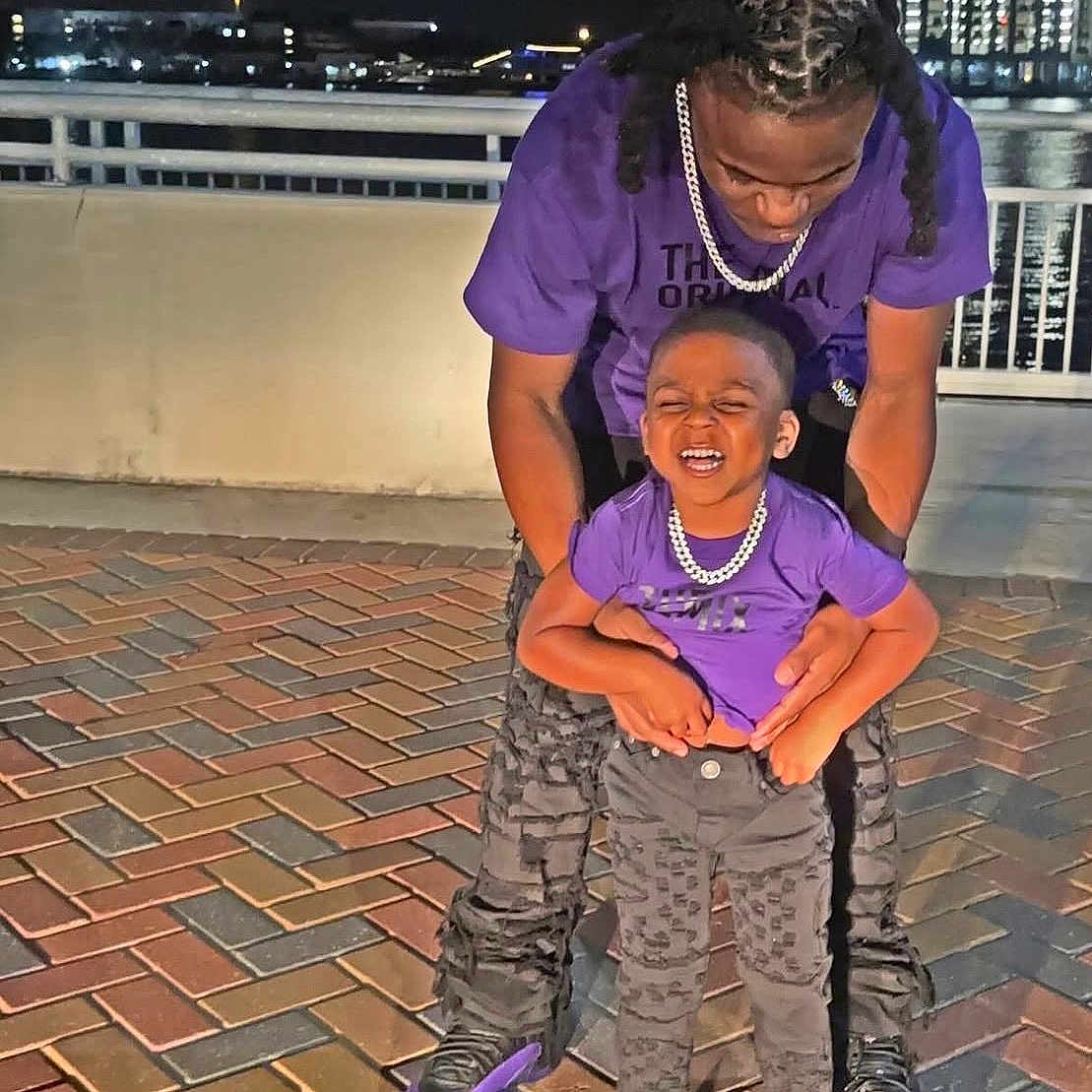 Josiah is registered to the contest to win money with this photo: adult, black_pants, brick, child, city_lights, family, fashion, happy, joy, matching_clothes, night, outdoor, pavement, person, playful, purple_shirt, railing, smile, sneakers, urban