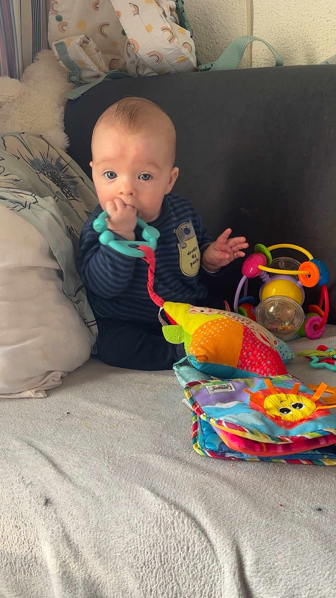 Jules participe au concours pour gagner de l'argent avec cette photo : baby, child, teething_ring, toy, colorful, couch, blanket, cushion, indoor, infant, playtime, cute, person, soft_toys, striped_clothing, hand, face, blue_eyes, sitting, home