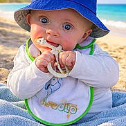 Ethan a rejoint le concours — aidez-le/la à gagner de superbes lots ! baby, beach, bib, blue_hat, bucket_hat, chewing, child, cute, giraffe, infant, ocean, outdoor, palm_tree, portrait, sand, smiling, sunny, teether, towel, wide_eyes