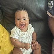 André participe au concours pour gagner de l'argent avec cette photo : baby, child, couch, cute, expression, face, fist, happy, human, indoor, infant, leather, person, portrait, sitting, skin, smiling, white_onesie, yellow_cushion, young