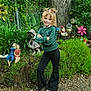 child, girl, green_sweater, black_pants, white_shoes, garden, plants, tree, statues, figurines, smile, pigtails, fence, outdoor, nature, gravel, playful, confident, portrait, casual_clothing
