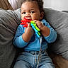 baby, child, infant, smiling, sitting, couch, indoor, portrait, curly_hair, blue_sweater, jeans, teething_toy, toy, colorful, happy, face, hands, home, cushion, photography
