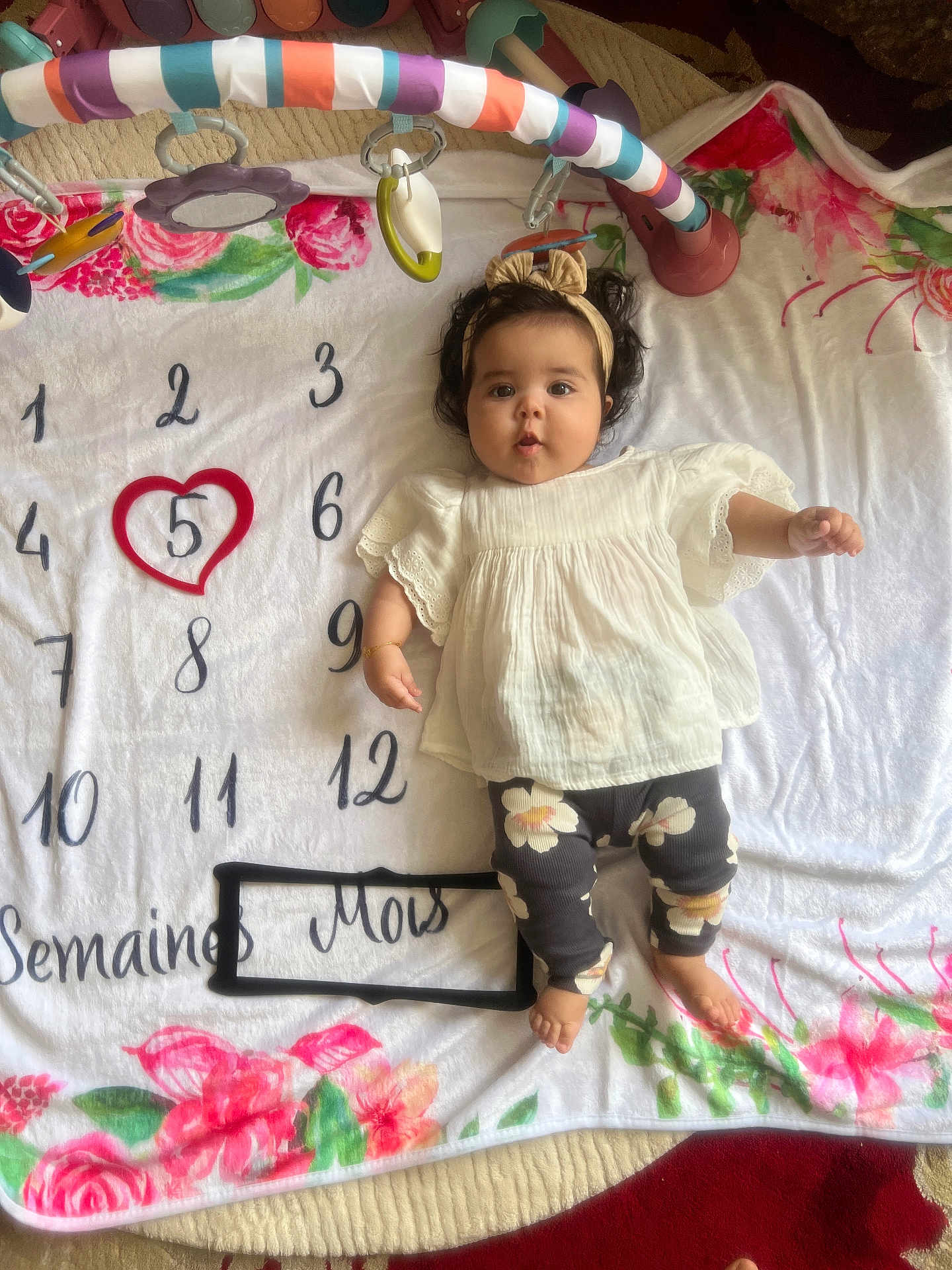 Ihssane a rejoint le concours — aidez-le/la à gagner de superbes lots ! baby, infant, milestone_blanket, headband, white_top, floral_leggings, toy_bar, colorful, numbers, heart, cute, lying_down, blanket, decorative, floor, child, young, indoors, portrait, feet