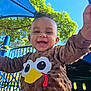 baby, blue_playground_equipment, brown_clothing, child, curly_hair, cute, daylight, face, fun, green_trees, happy, joy, metal_play_structure, outdoor, park, playground, smiling, sunny_day, toddler, turkey_sweater