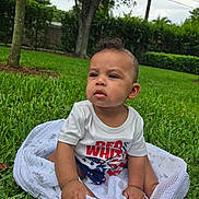 Zayden is registered to the contest to win money with this photo: baby, grass, blanket, outdoor, child, shirt, patriotic, red_white_blue, sitting, greenery, tree, yard, footwear, curly_hair, expression, nature, daylight, person, casual, peaceful