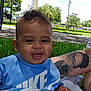 baby, child, smiling, blue_shirt, nike, grass, outdoor, tattoo, arm, blanket, park, trees, sitting, happy, person, playful, sunny, daytime, face, clothing