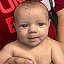 Carter is registered to the contest to win money with this photo: baby, infant, child, face, blue_eyes, skin, hand, adult_hand, holding, red_shirt, text_on_shirt, portrait, close_up, human, person, expression, curious, indoor, young, cute