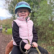 Kamilia participe au concours pour gagner de l'argent avec cette photo : child, helmet, pony, horseback_riding, outdoor, smile, vest, reins, nature, trail, autumn, greenery, happy, person, animal, animal_riding, young_child, casual_clothing, daytime, forest