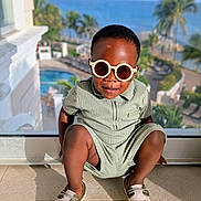 Temi is registered to the contest to win money with this photo: toddler, child, sunglasses, romper, shoes, indoor, glass_window, sea, palm_trees, sunlight, smiling, fashion, pose, floor, resort, vacation, outdoor_view, shadow, happy, portrait