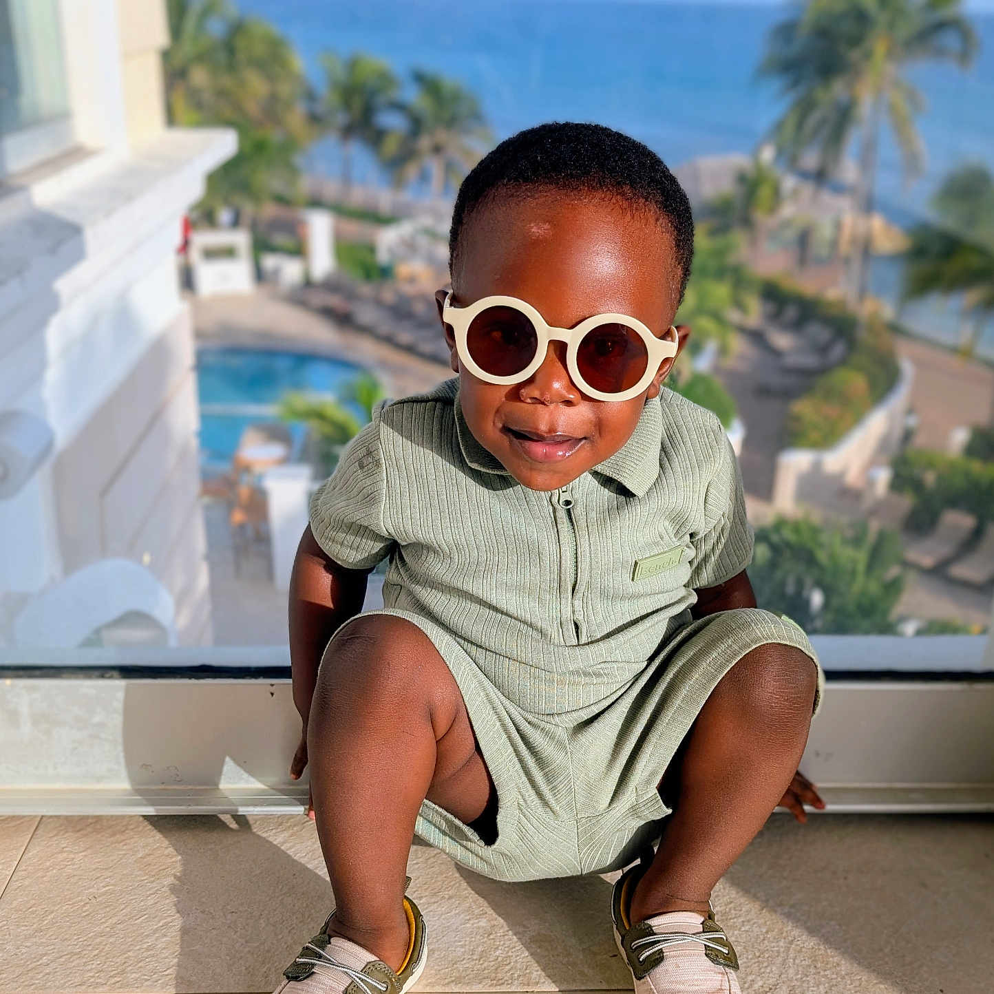 Temi is registered to the contest to win money with this photo: child, fashion, floor, glass_window, happy, indoor, outdoor_view, palm_trees, portrait, pose, resort, romper, sea, shadow, shoes, smiling, sunglasses, sunlight, toddler, vacation