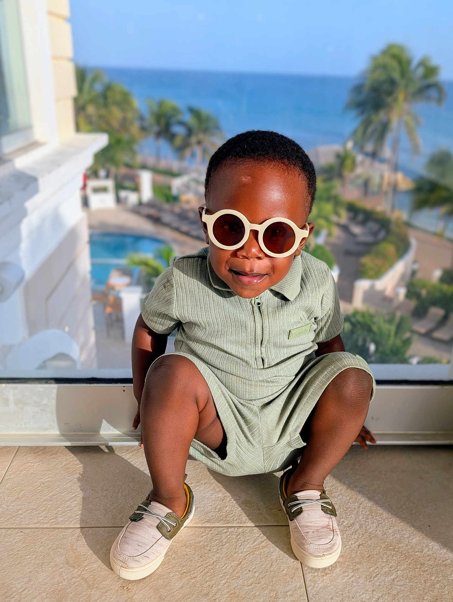 Temi is registered to the contest to win money with this photo: toddler, child, sunglasses, romper, shoes, indoor, glass_window, sea, palm_trees, sunlight, smiling, fashion, pose, floor, resort, vacation, outdoor_view, shadow, happy, portrait