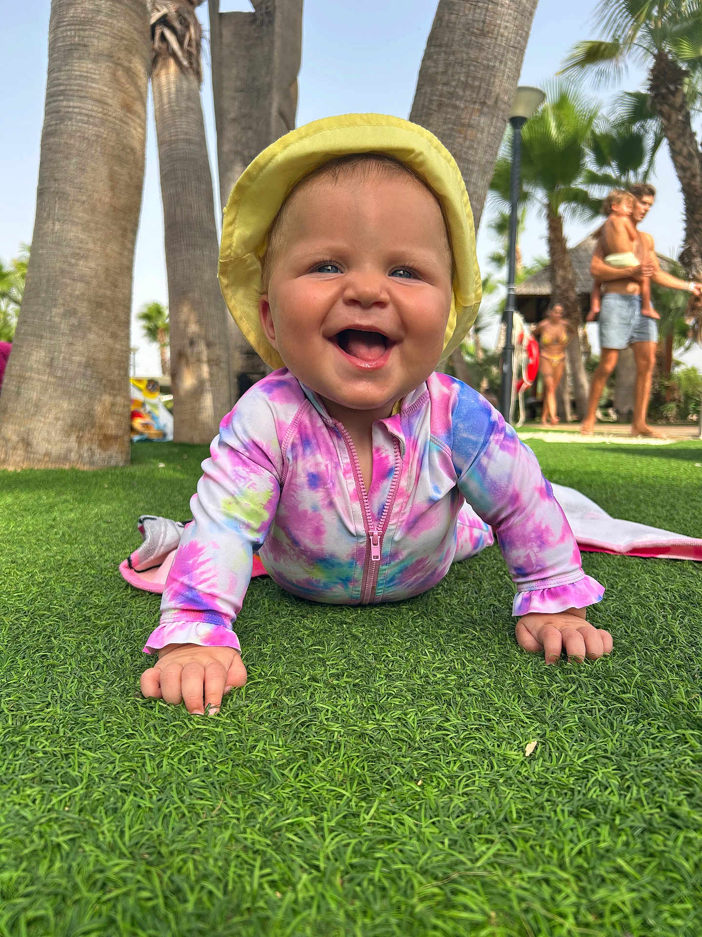 Millie joined the competition — help win amazing prizes! baby, child, smiling, crawling, grass, outdoor, sunhat, tie_dye_clothing, palm_trees, people, summer, vacation, happy, nature, family, playful, sunny, blue_sky, greenery, daytime