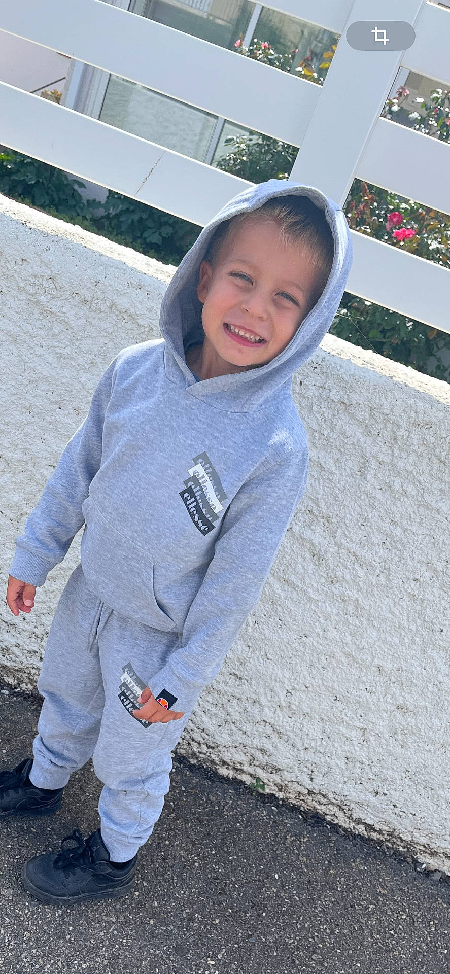 Nino a rejoint le concours — aidez-le/la à gagner de superbes lots ! black_sneakers, boy, casual_clothing, child, daylight, fashion, flowers, happy, hoodie, outdoor, pavement, person, portrait, smiling, standing, street, sweatpants, white_fence, white_wall, young