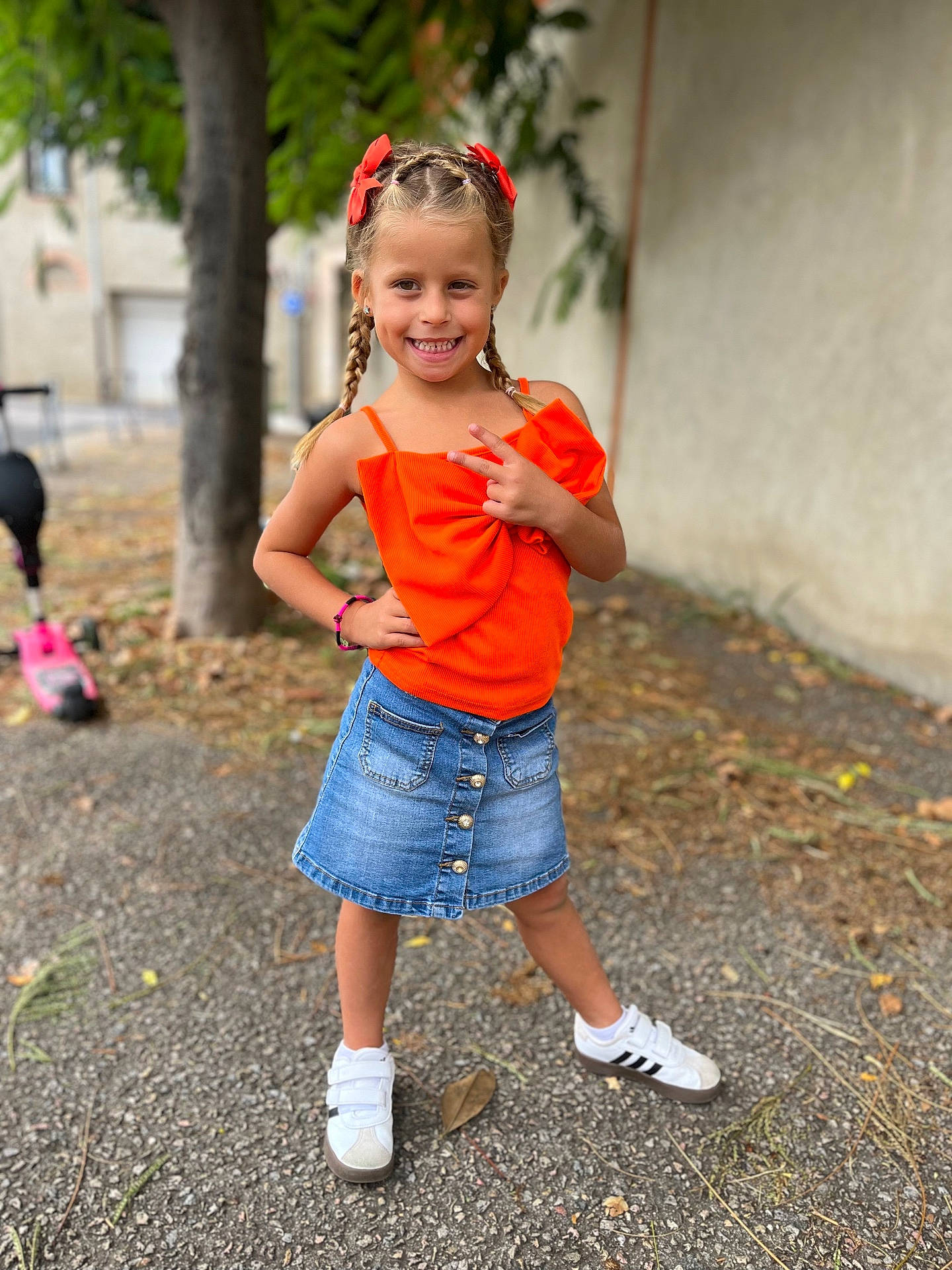 Giulia participe au concours pour gagner de l'argent avec cette photo : background_blur, braids, casual_clothing, child, daylight, denim_skirt, fashion, girl, happy, orange_top, outdoor, pavement, peace_sign, person, playful, posing, red_bows, smiling, sneakers, tree