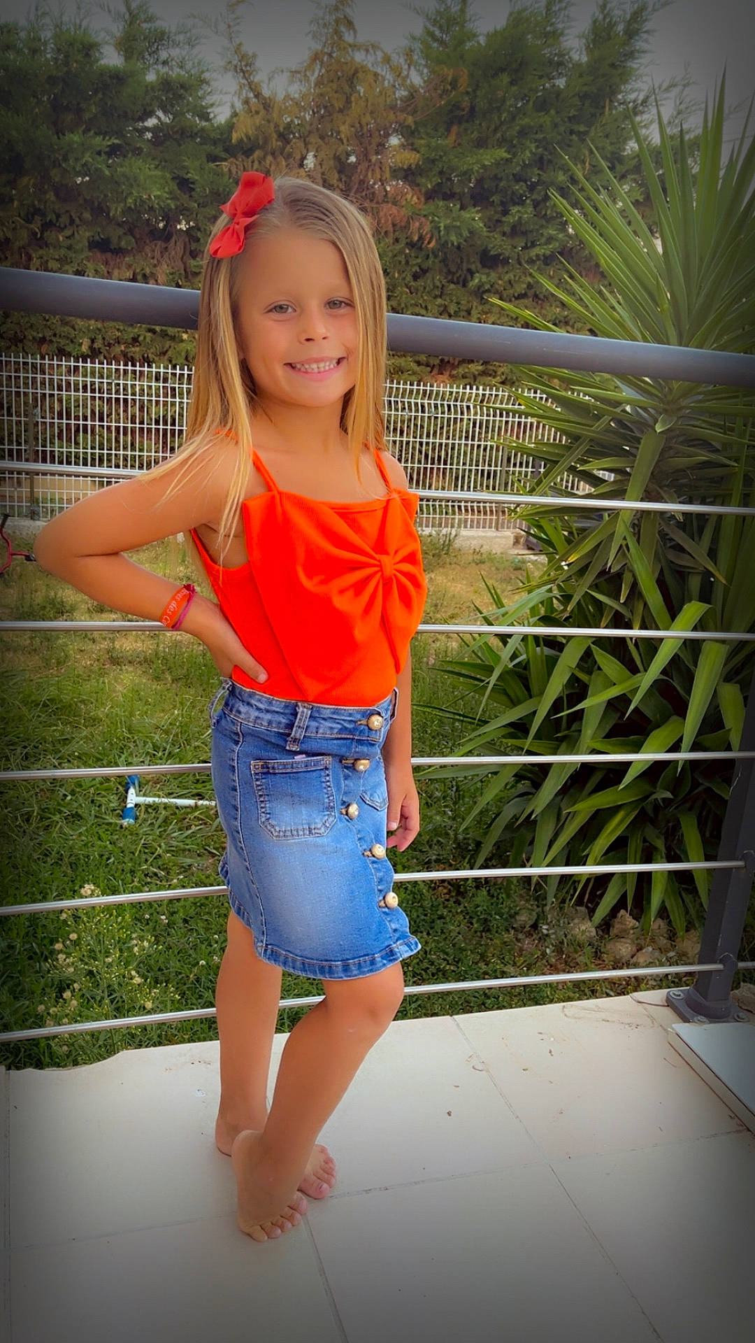 Giulia a rejoint le concours — aidez-le/la à gagner de superbes lots ! barefoot, blonde_hair, casual_clothing, child, daylight, denim_skirt, fence, girl, grass, greenery, happy, metal_railing, nature, orange_top, outdoor, portrait, posing, smiling, summer, young