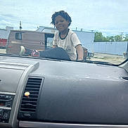 Ta'Ron is registered to the contest to win money with this photo: child, smiling, car_dashboard, car_interior, hood, outdoor, van, building, sky, cloudy, person, casual_clothing, dreadlocks, urban, parking_lot, vehicle, young, happy, daylight, relaxed