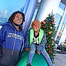 child, man, outdoor, christmas_tree, holiday_decorations, large_ornament, building, glass_windows, metallic_columns, casual_clothing, hat, jeans, sneakers, smiling, posing, urban, festive, winter_clothes, daylight, sidewalk