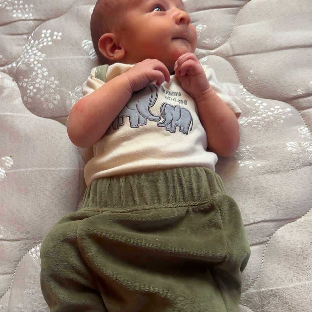 Daxsyn is registered to the contest to win money with this photo: baby, child, clothing, curious, cute, elephant_design, face, foot, green_pants, hand, indoor, infant, laying_down, mattress, newborn, pattern, person, remote_control, sleepwear, soft_texture