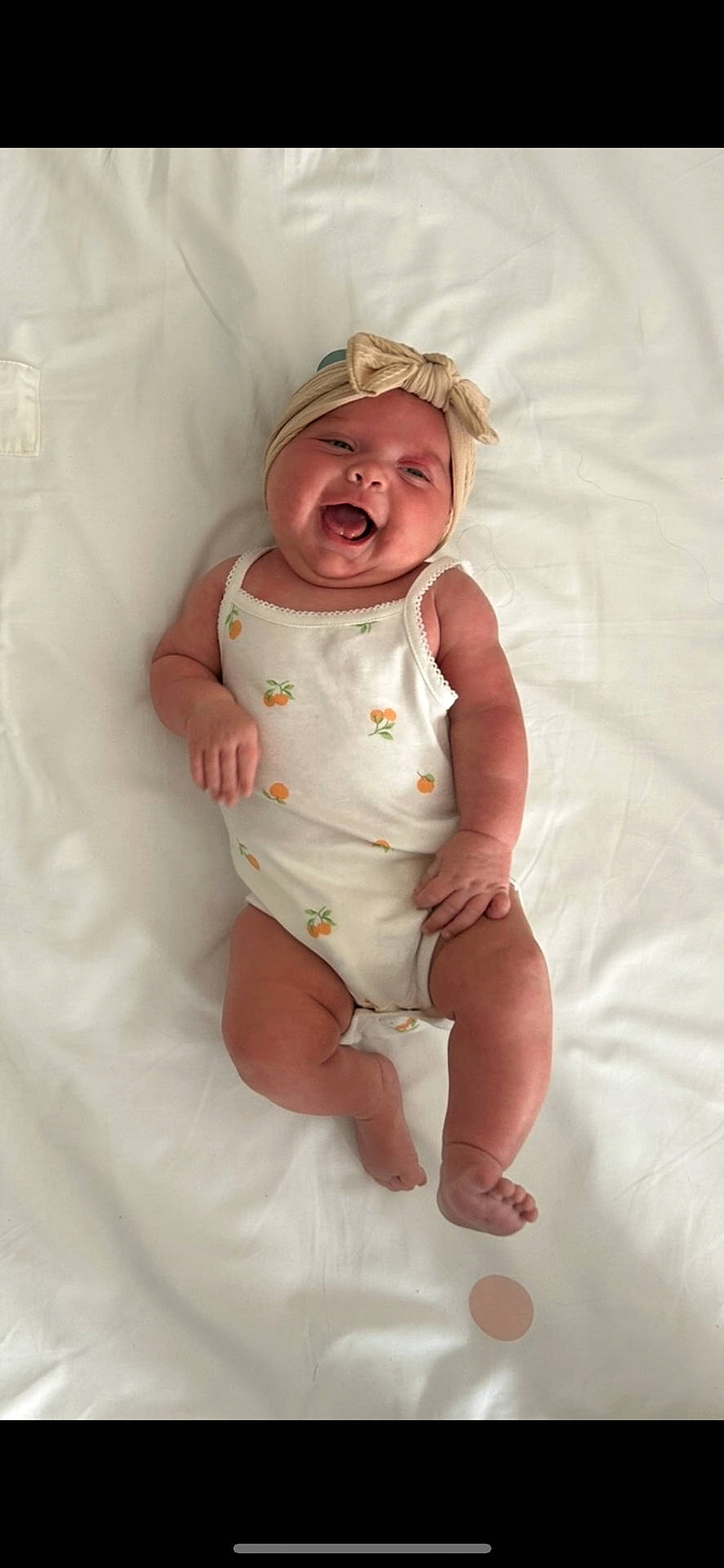 apparel, baby, child, cute, face, happy, headband, human, indoors, infant, lying_down, onesie, patterned_clothing, portrait, skin, sleepwear, smiling, soft_lighting, tongue_out, white_bed