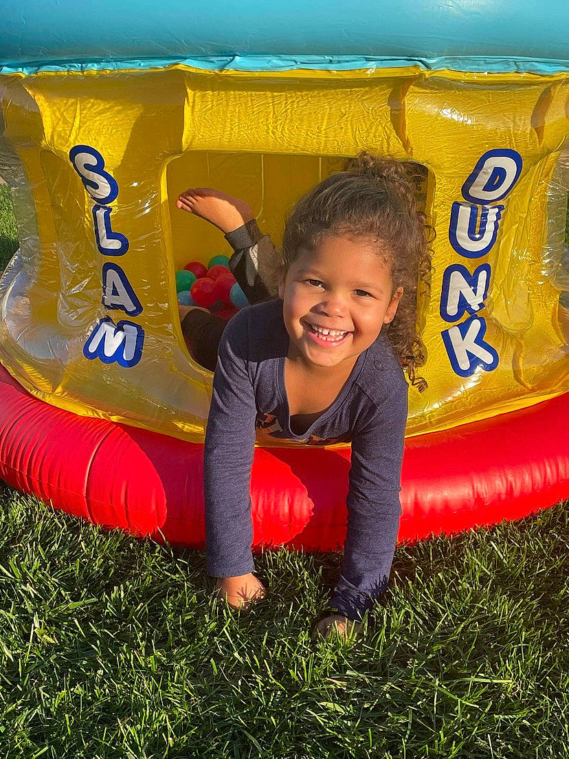Kali is registered to the contest to win money with this photo: aircraft, bounce_house, child, chute, competition_event, fun, games, grass, happy, inflatable, joy, leisure, outdoor_play_equipment, people_in_nature, person, plant, play, playground, recreation, smile
