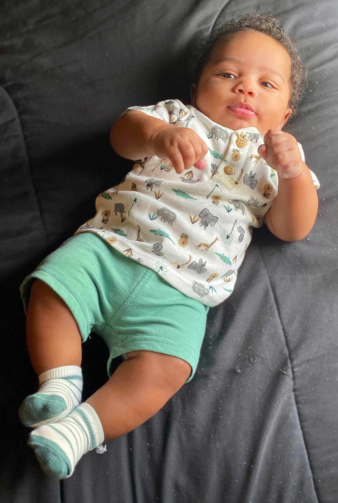 Quadarius is registered to the contest to win money with this photo: baby, infant, child, clothing, shirt, shorts, socks, animal_print, curly_hair, skin, face, eyes, hands, legs, footwear, bed, quilt, relaxing, indoors, portrait
