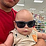 Noah joined the competition — help win amazing prizes! baby, child, sunglasses, portrait, indoor, store, retail, shopping, adult, parent, holding, infant, clothing, red_shirt, toucan_pattern, serious, fashion, selfie, face, shelving