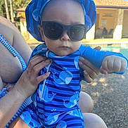 Noah is registered to the contest to win money with this photo: baby, child, infant, sunglasses, blue_outfit, swimwear, poolside, outdoor, sunny, person, face, portrait, being_held, adult_hands, whale_pattern, hat, serious_expression, fist, palm_trees, house