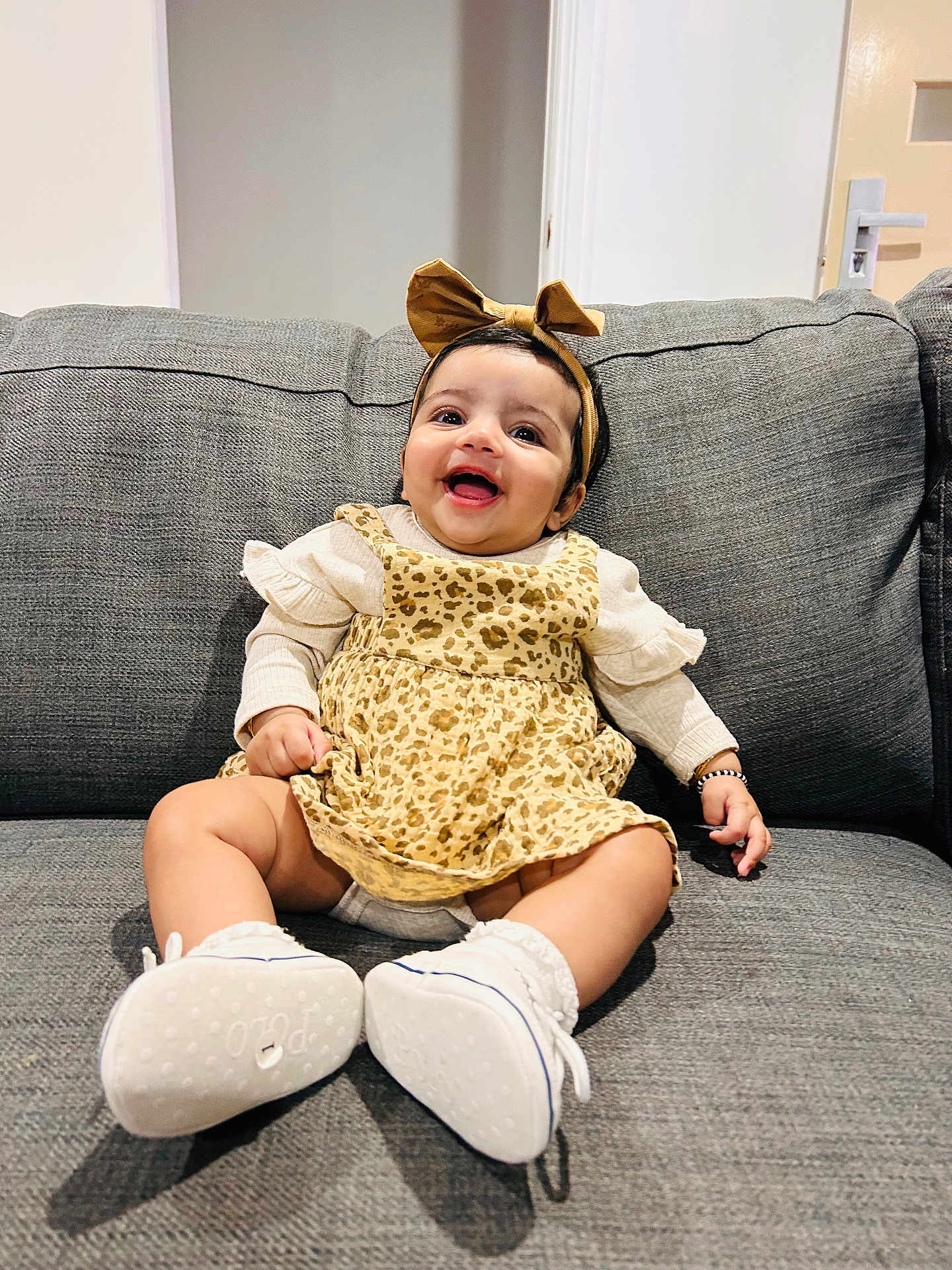 Kiaana joined the competition — help win amazing prizes! baby, infant, smile, couch, sofa, headband, bow, leopard_print, dress, white_shoes, socks, sitting, happy, portrait, living_room, furniture, door, wall, eyes, hands