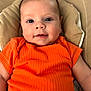 baby, child, infant, orange_shirt, ribbed_clothing, cushion, beige, face, eyes, smile, skin, head, person, portrait, closeup, comfortable, cute, young, lying_down, indoor