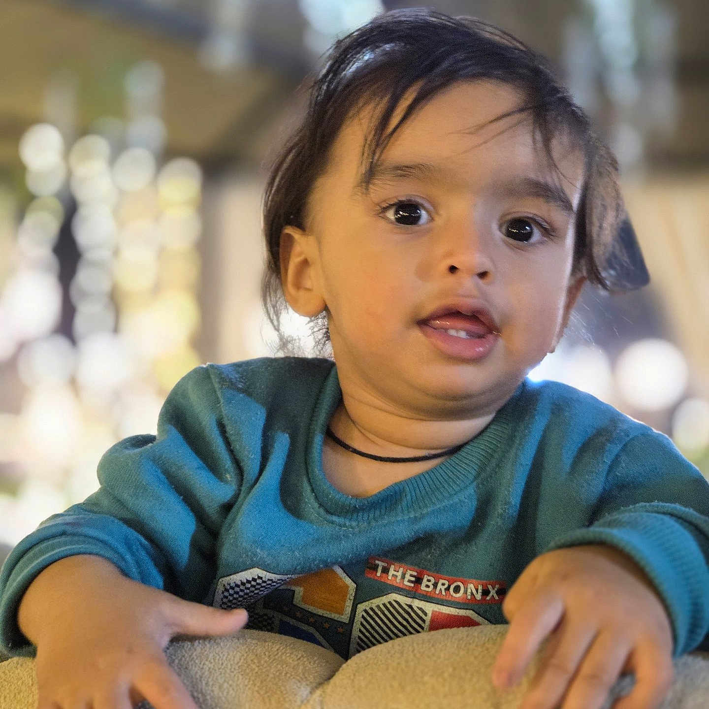 Vihaan a rejoint le concours — aidez-le/la à gagner de superbes lots ! child, toddler, face, hands, sweater, cushion, indoor, portrait, curly_hair, expression, young, person, casual_clothing, soft_lighting, blurred_background, warm_colors, cute, looking_at_camera, close_up, cozy