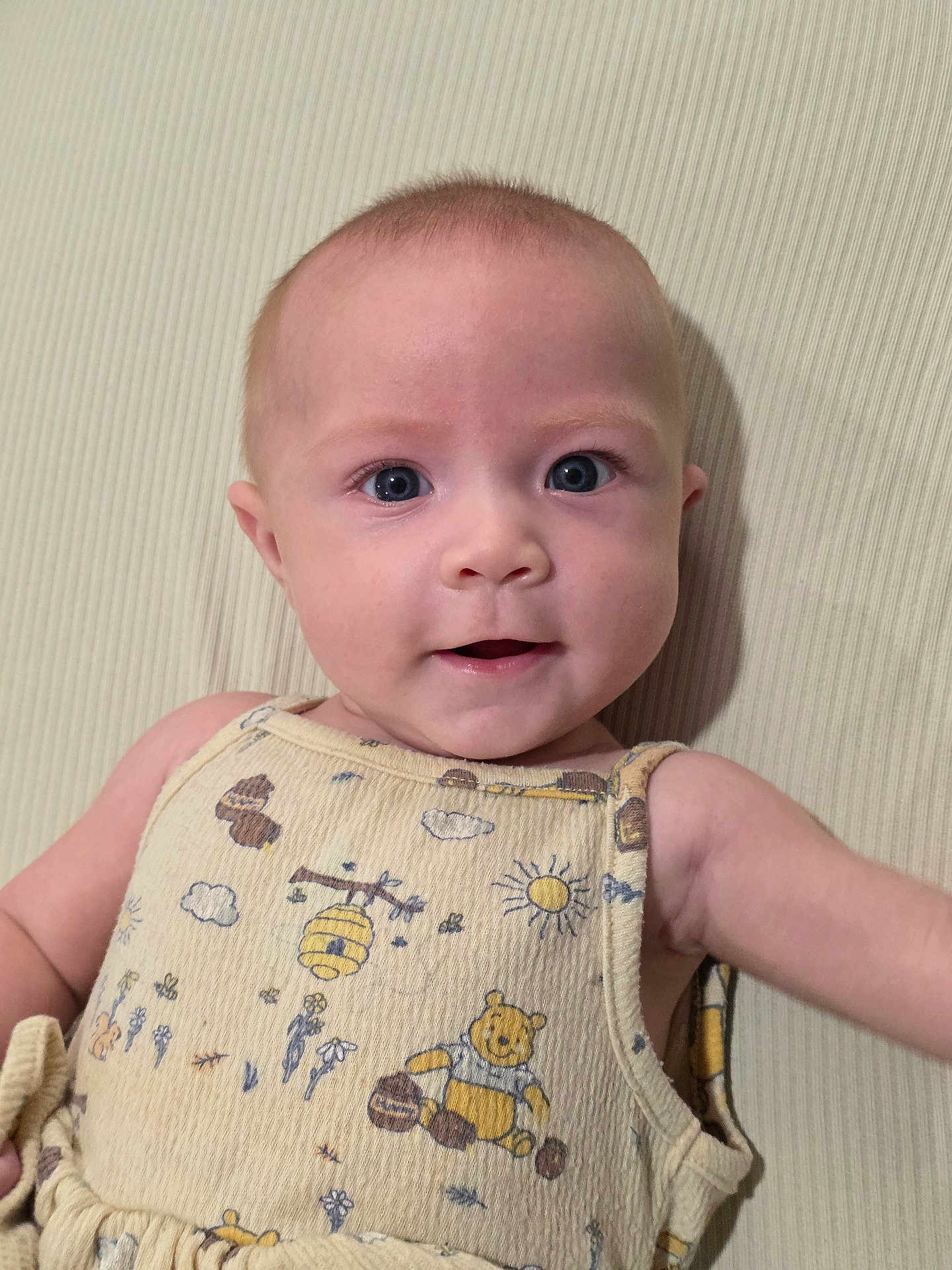 Alexis joined the competition — help win amazing prizes! baby, infant, child, face, blue_eyes, smile, onesie, winnie_the_pooh, patterned_clothing, portrait, closeup, cheeks, eyelashes, arm, skin, plaid_background, textile, soft_lighting, indoor, nursery