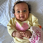 Jenaiah a rejoint le concours — aidez-le/la à gagner de superbes lots ! baby, infant, smiling, blanket, bed, yellow_sweater, pink_heart, bow, curly_hair, bracelet, plush_toy, hands, face, portrait, indoor, soft_texture, cute, lying_down, cheeks, natural_light