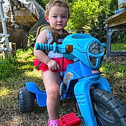 Sabrina joined the competition — help win amazing prizes! child, toddler, outdoor, backyard, ride_on_toy, motorcycle_toy, blue_vehicle, sitting, portrait, face, serious_expression, brown_hair, summer, sunlight, grass, trees, shed, water_barrel, pink_clothing, playtime