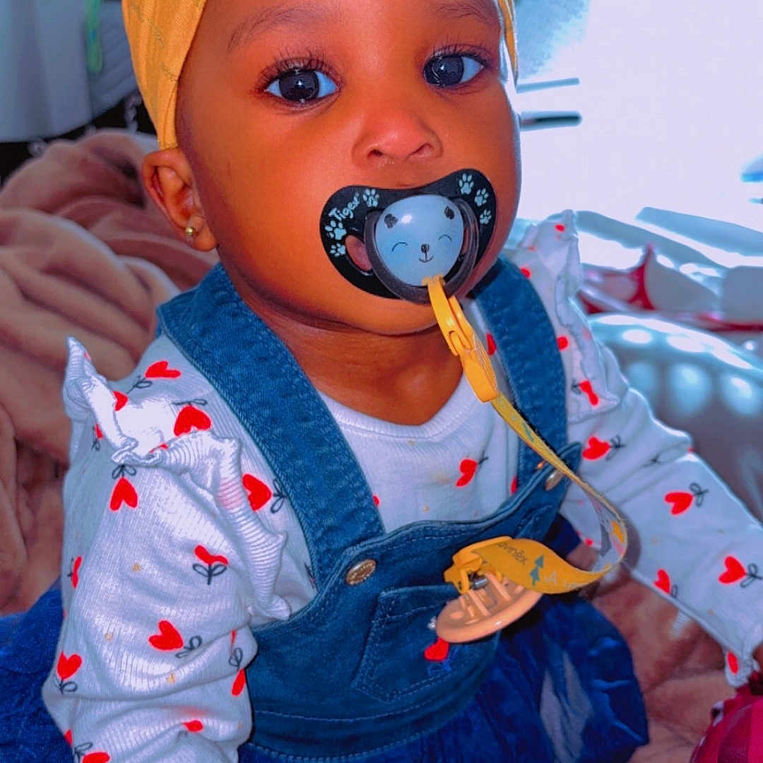Fatoumata a rejoint le concours — aidez-le/la à gagner de superbes lots ! baby, baby_product, blanket, child, clothing, colorful, cute, denim, dress, eyes, face, hand, headwrap, heart_emoji, indoor, pacifier, person, room, sitting, toy